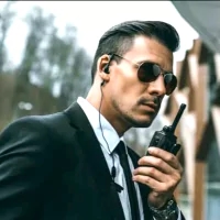 the image is a man in a suit and sunglasses holding a walkie talkie